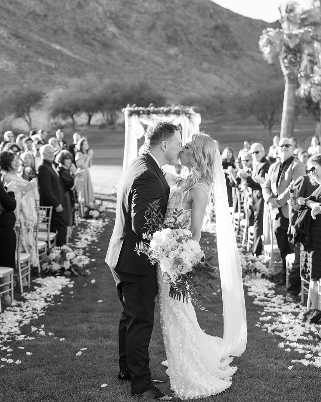 Black and white photo of a bride and groom kissing at the altar during an outdoor Las Vegas wedding ceremony, bride holding a white rose bouquet