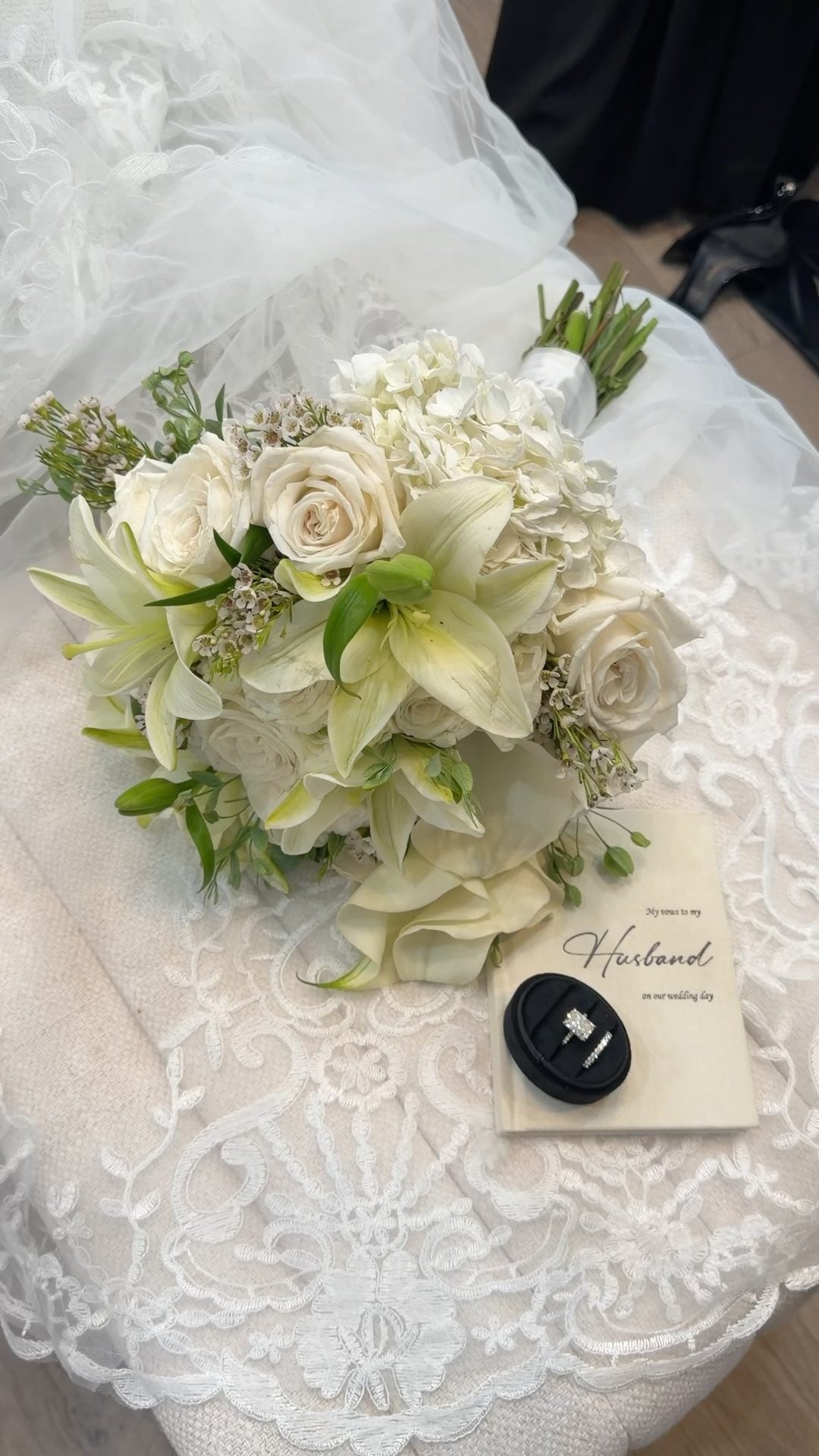 White bridal bouquet of lilies, roses, hydrangea and calla lilies resting on lace beside the wedding rings and a vow booklet